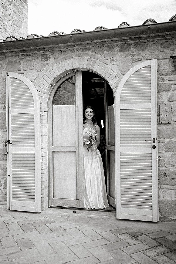 Bridal portrait of a smiling bride holding bouquet in a strapless satin dress, standing in an arched doorway by a stone wall
