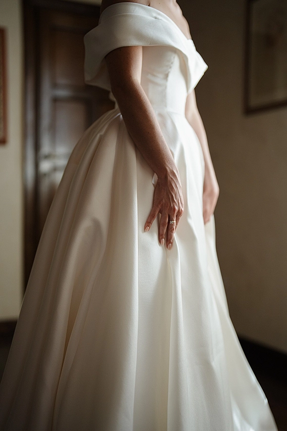 Wedding dress with off the shoulder wedding dress neckline in satin, full skirt shown in indoor doorway as bride displays ring and nude manicure