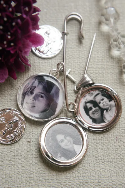 Photo locket charms with wedding photo charm inserts pinned on a safety pin, silver lockets and crystal beads beside burgundy flowers on textured fabric