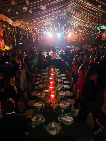 Reception tablescape with a long banquet table wedding setup, greenery garland, red lamps, and hanging glass votives under a draped tent ceiling at night
