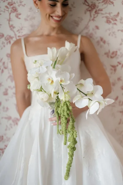 Bridal portrait of a bride holding bouquet of white orchid blooms in a strapless wedding dress, pearl earrings, against floral wallpaper