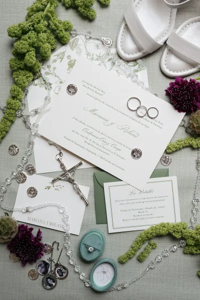Invitation suite with botanical wedding invitations, envelope, rosary and silver coins, ring box and white sandals on gray linen with purple flowers