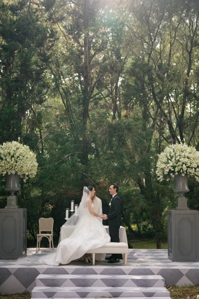 Wedding vows as bride and groom hold hands at an outdoor wedding ceremony, bride’s veil flowing beside candlelit altar in sunlit garden