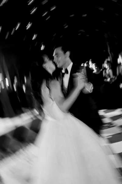 First dance as bride twirls in her wedding dress with groom in tuxedo, dramatic lighting in a dark reception space with guests watching