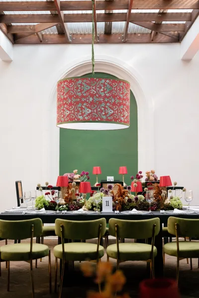 Reception tablescape with long banquet table decor, red lamps, floral centerpieces and grapes, set under drum pendant light in arched hall