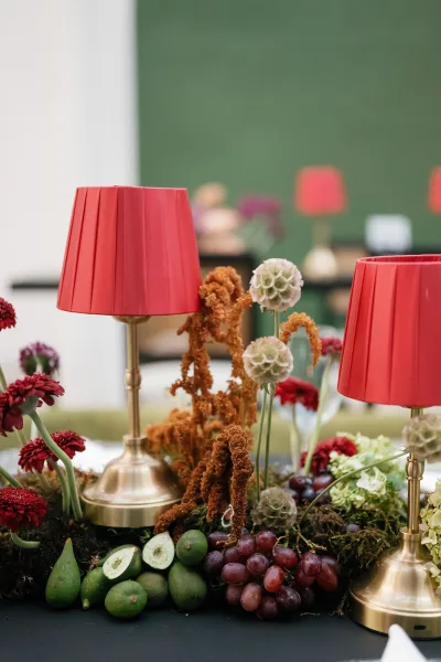 Reception tablescape with a wedding table centerpiece of red lamps on brass bases, moss runner with grapes and figs, against a green wall