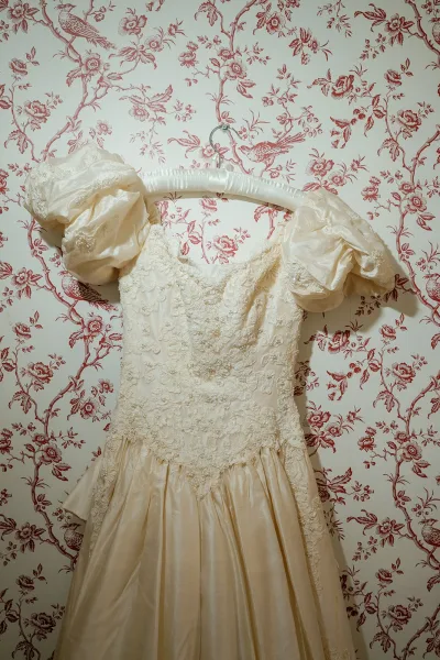 Wedding dress with lace bodice and beading, satin skirt and puff sleeves hanging on a padded hanger against floral bird wallpaper backdrop