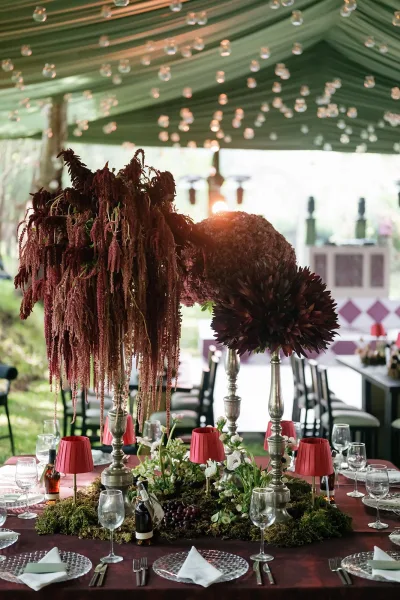 Reception tablescape with tall wedding centerpieces, silver candlesticks, and burgundy tablecloth under draped tent ceiling and string lights outdoors