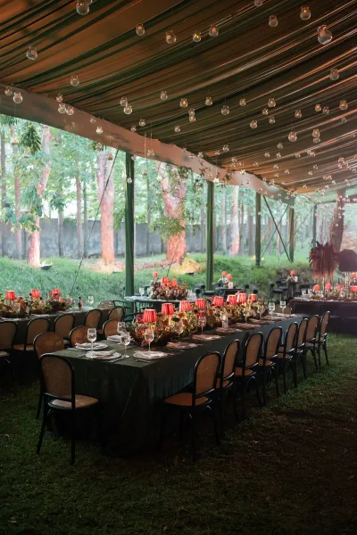 Reception tablescape with long banquet table setup, dark linens, floral garland, red lamps, candles and hanging glass votives under a tent canopy