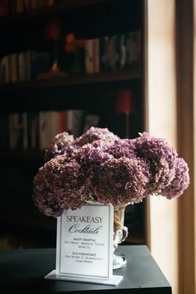 Wedding bar menu and signature cocktail menu displayed in a clear holder beside hydrangeas in a vase, set by a bookshelf in window light