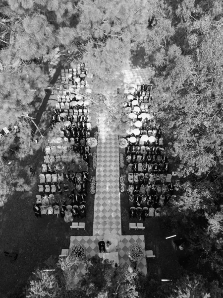 Ceremony setup for an outdoor wedding ceremony with checkered aisle runner, chair rows, floral markers, and white parasols under trees