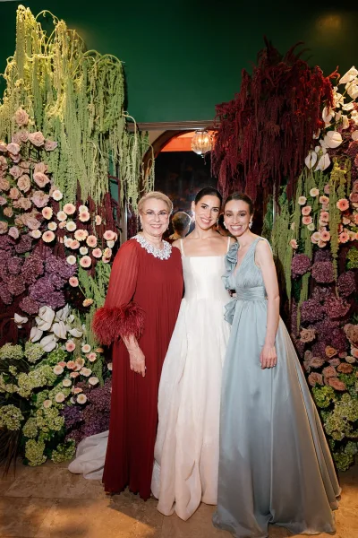 Bridal party portrait with bride in a strapless corset wedding dress beside mother and bridesmaid before a floral wall and arched doorway indoors.