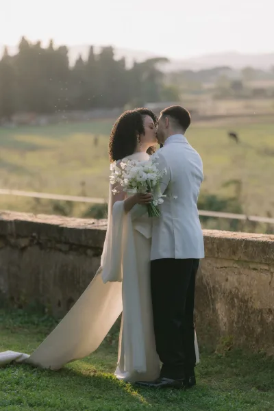 Wedding kiss portrait of bride and groom kissing as she holds a white bouquet, with veil flowing by a stone wall at sunset