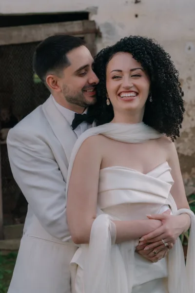 Couple portrait of bride and groom embrace, groom hugging her from behind as she laughs by a rustic stone wall and doorway