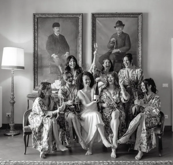 Bridesmaid getting ready in floral robes holding champagne flutes beside a bride in slip dress, laughing in a bright indoor suite