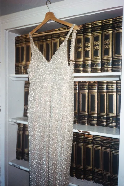 Wedding dress hanging on a wooden hanger, featuring beaded fabric and spaghetti straps, set against a bookshelf of vintage books