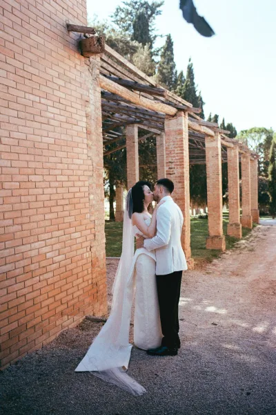 Wedding kiss portrait of bride and groom kissing in side profile, her long veil train flowing by brick columns and pergola beams outdoors