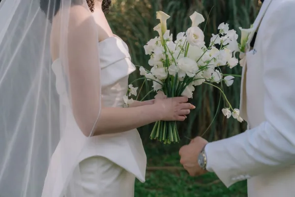 Wedding bouquet of white calla lilies held by bride in strapless dress beside groom in suit with wristwatch, garden greenery behind