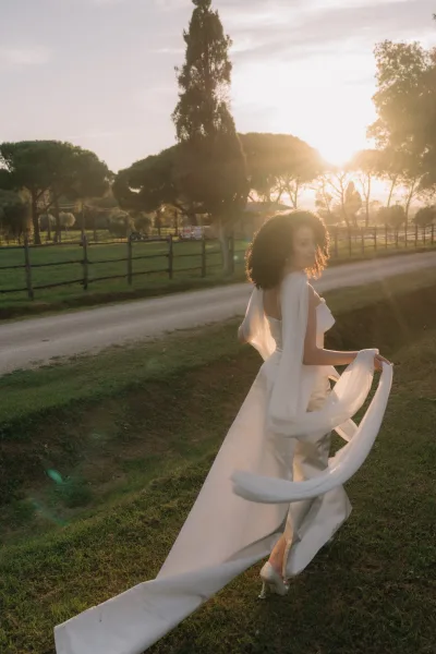 Bridal portrait of a bride in a long sleeve open-back wedding dress walking away on a dirt road at sunset, looking back in a grassy field