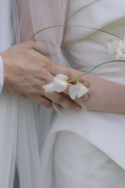 Wedding hands clasped beneath a bridal veil, resting on white dress fabric with a white flower and green stems against a soft neutral backdrop