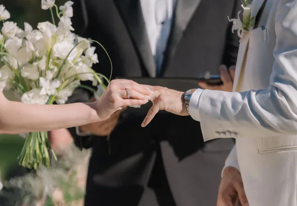 Wedding ring exchange close up of bride sliding a diamond band onto groom’s hand, showing her manicure, his watch, and white bouquet at the ceremony