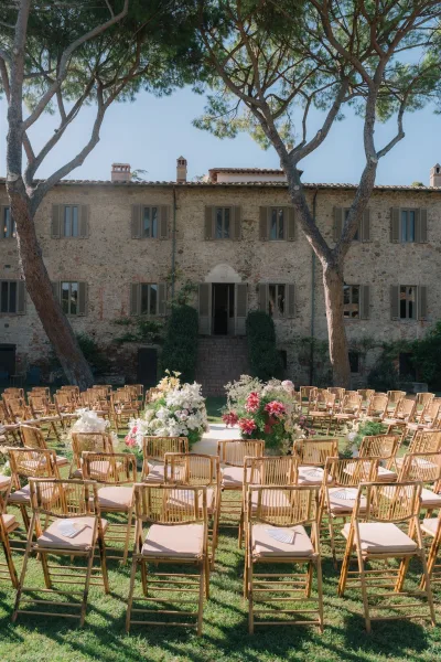 Ceremony setup with outdoor wedding ceremony seating in a circle of wood chairs around a flowered platform on a lawn by a stone villa under blue sky
