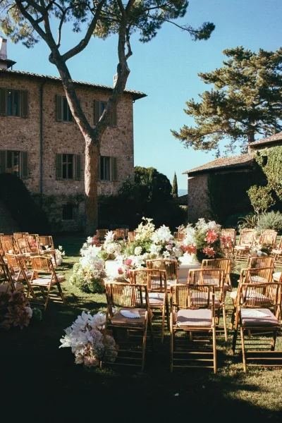 Outdoor ceremony setup with garden wedding ceremony seating in a circle, wood chairs, white aisle runner, and pastel flowers by a stone villa