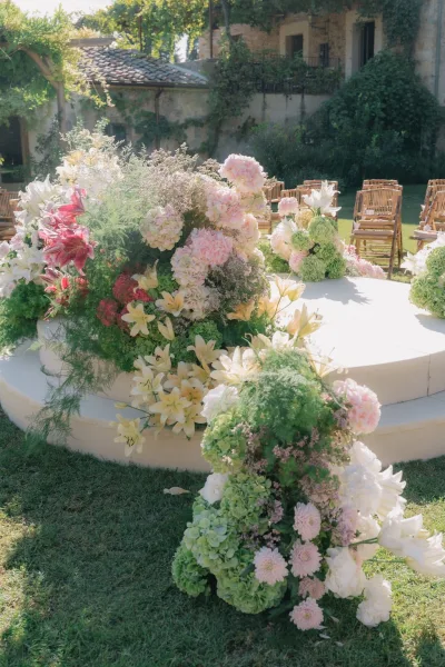 Ceremony altar decor on a white circular platform with pastel hydrangeas and lilies, flanked by wooden chairs on a sunny lawn by a vine-covered stone building