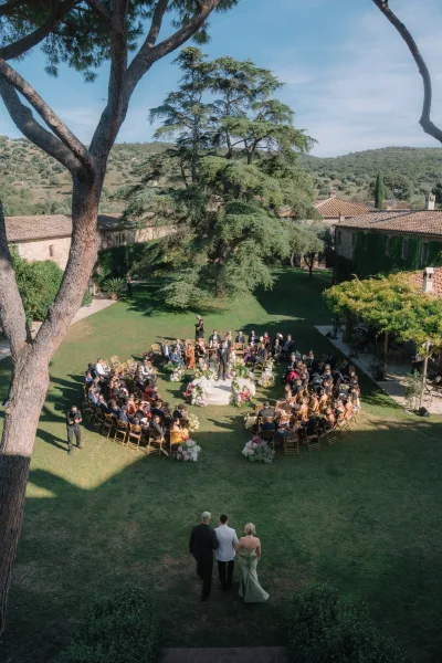 Wedding processional at an outdoor wedding ceremony with gold Chiavari chairs circling a round platform, aisle florals on a sunny lawn under trees