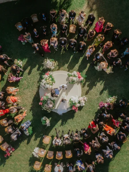 Outdoor wedding ceremony seen from above, with guests seated in a circle around floral arrangements and a veiled bride on a sunlit lawn