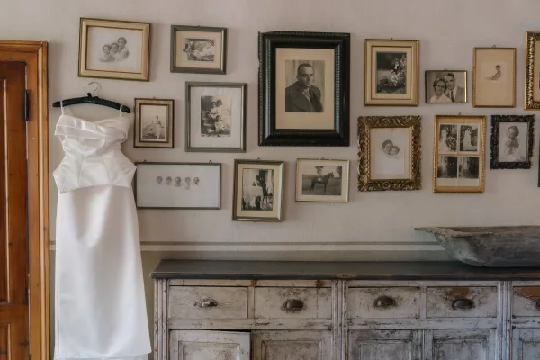 Wedding dress hanging on a hanger beside a vintage dresser, framed family photo gallery wall behind on a white room wall