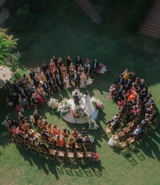 Outdoor wedding ceremony with circular wedding ceremony seating, bride and groom at a floral arch amid wooden chairs on a garden lawn from above