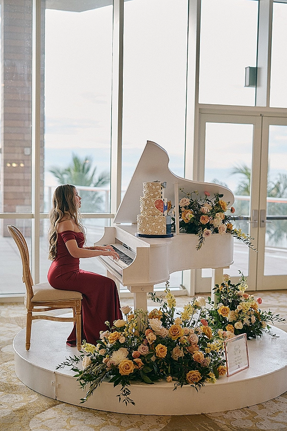 Wedding pianist in an off-shoulder burgundy dress playing a white grand piano with cake and florals by ocean-view windows