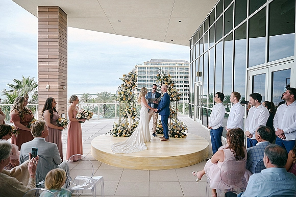 Wedding ceremony with bride and groom at a floral arch on an outdoor terrace, clear acrylic chairs, palm trees and ocean view under clouds