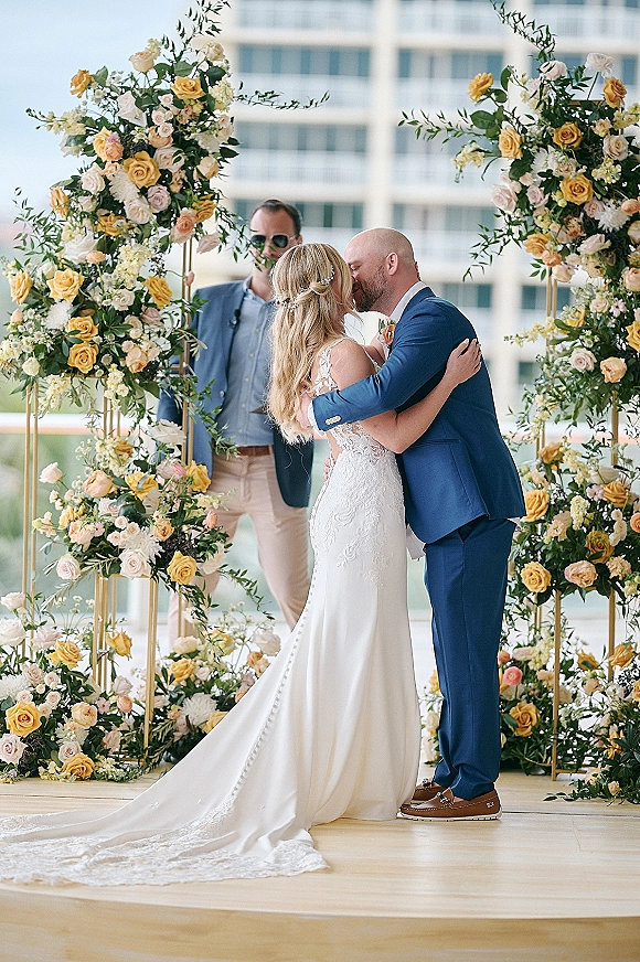 Wedding kiss under a rose floral arch on an outdoor balcony by the water, bride in lace gown and veil, groom in blue suit