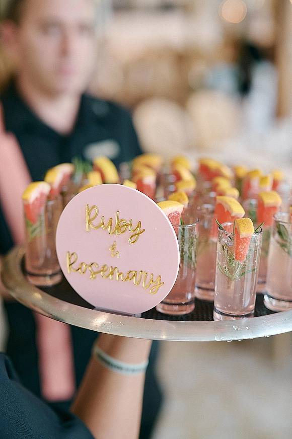 Wedding welcome drinks with signature wedding cocktails on a cocktail tray, shot glasses topped with grapefruit and rosemary beside a round gold sign at an indoor bar