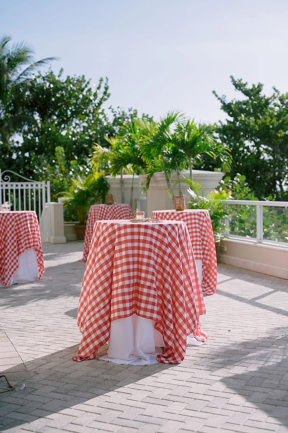Cocktail hour tables with red gingham tablecloths, round high tops and simple centerpieces on a brick patio with palm trees and blue sky