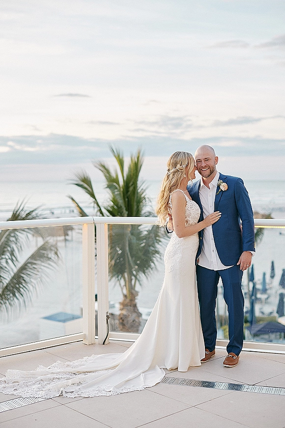 Couple portrait of bride and groom embracing on a balcony with ocean view, her lace dress long train and hair vine, palm trees behind