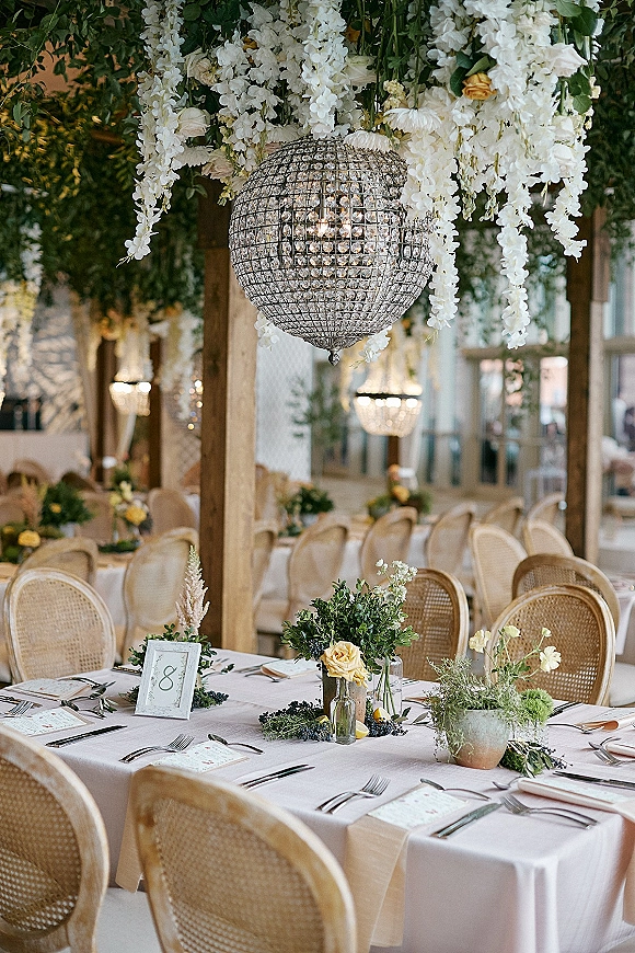 Reception tablescape with wedding table setting on white linen, yellow rose bud vases and greenery garland beneath a crystal chandelier in a timber-beam hall