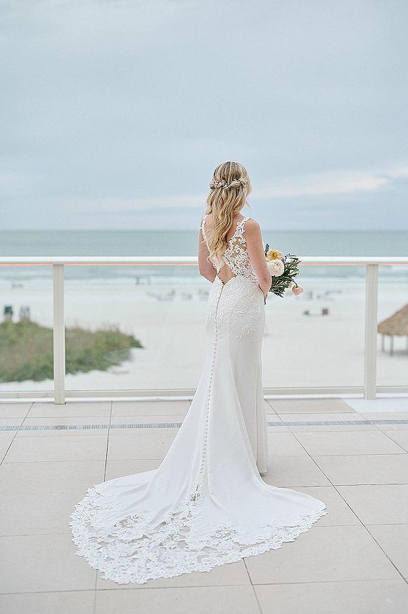 Bridal portrait of a bride from behind in an open back lace wedding dress with long train, holding bouquet on a balcony with ocean view