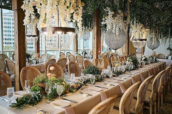 Reception tablescape with a long table wedding reception setup featuring greenery garland, candles, cane-back chairs, and floral ceiling under chandeliers