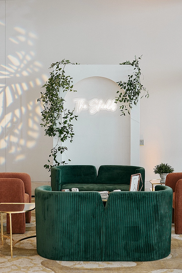 Wedding lounge area with a green velvet sofa, gold side tables, and neon sign against an arch backdrop with greenery garland indoors