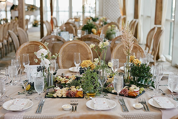 Reception tablescape with wedding table decor, yellow rose bud vases and greenery garland, candles and charcuterie set in a sunlit room