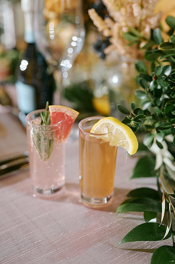 Signature wedding cocktails with grapefruit garnish and rosemary sprig in cocktail glasses on a reception table with greenery runner and soft bokeh lighting