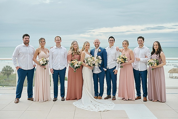Wedding party portrait with bride and groom holding bouquets on a balcony terrace with glass railing, ocean and cloudy sky behind