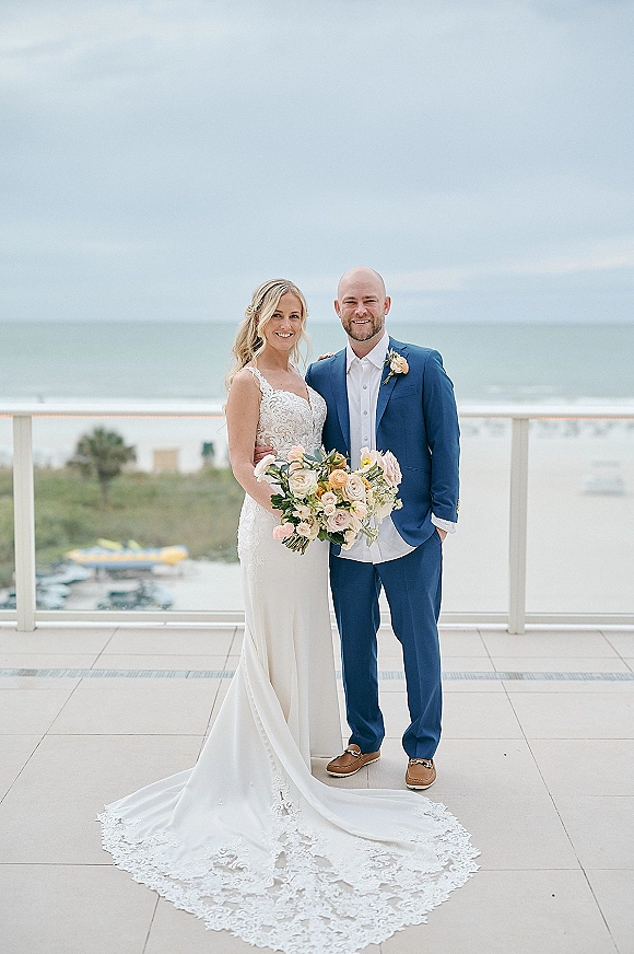 Couple portrait of bride holding bouquet beside groom in blue suit, her lace dress train flowing on a tiled oceanfront balcony under cloudy sky