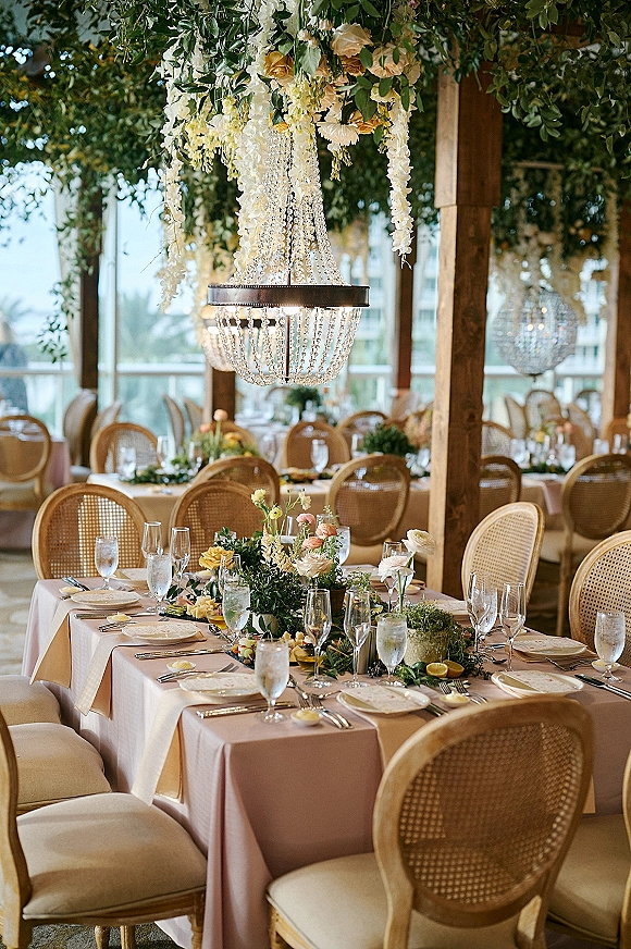 Reception tablescape with blush wedding table decor, lush floral centerpieces and greenery garlands beneath crystal chandeliers in a wood-beamed room