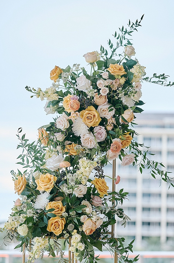 Wedding floral arch with roses and mixed greenery on a modern frame, set against open sky and a sleek modern building backdrop