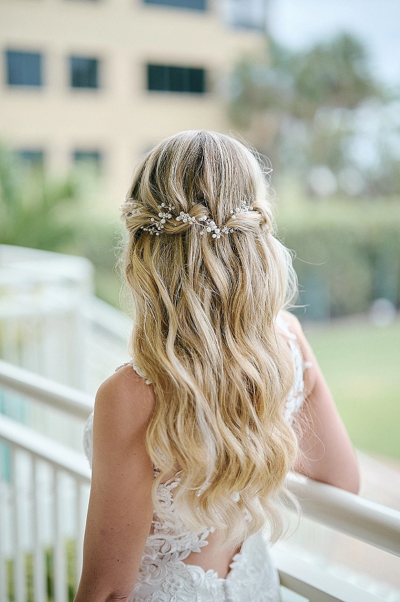 Bridal hairstyle with half up half down wedding hair, soft blonde waves and pearl hair vine, shown from behind on a balcony with trees