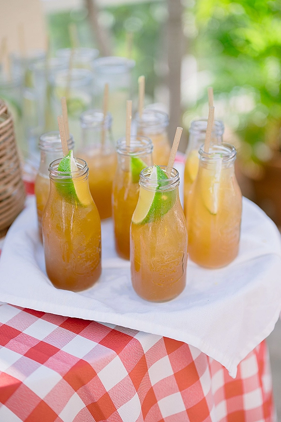 Wedding drink station with signature drink bottles, citrus wedges and wooden stir sticks on a white tray with wicker basket outdoors